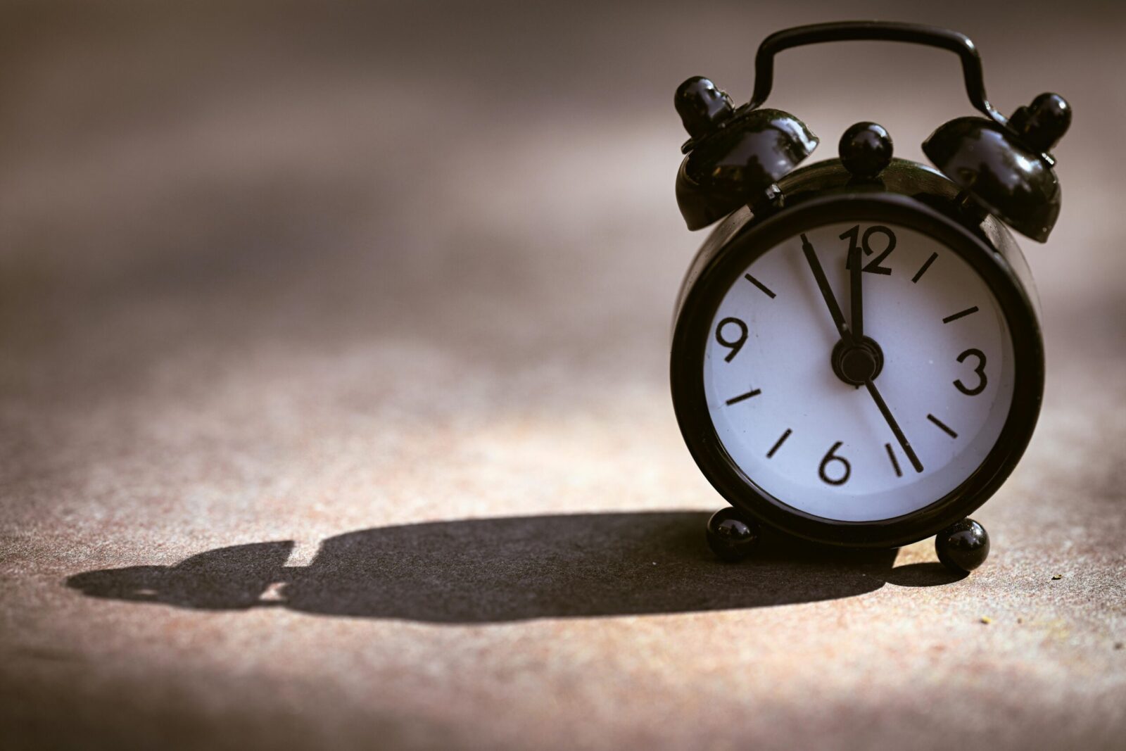 A classic black alarm clock showing five minutes to twelve on a textured surface with dramatic shadow.