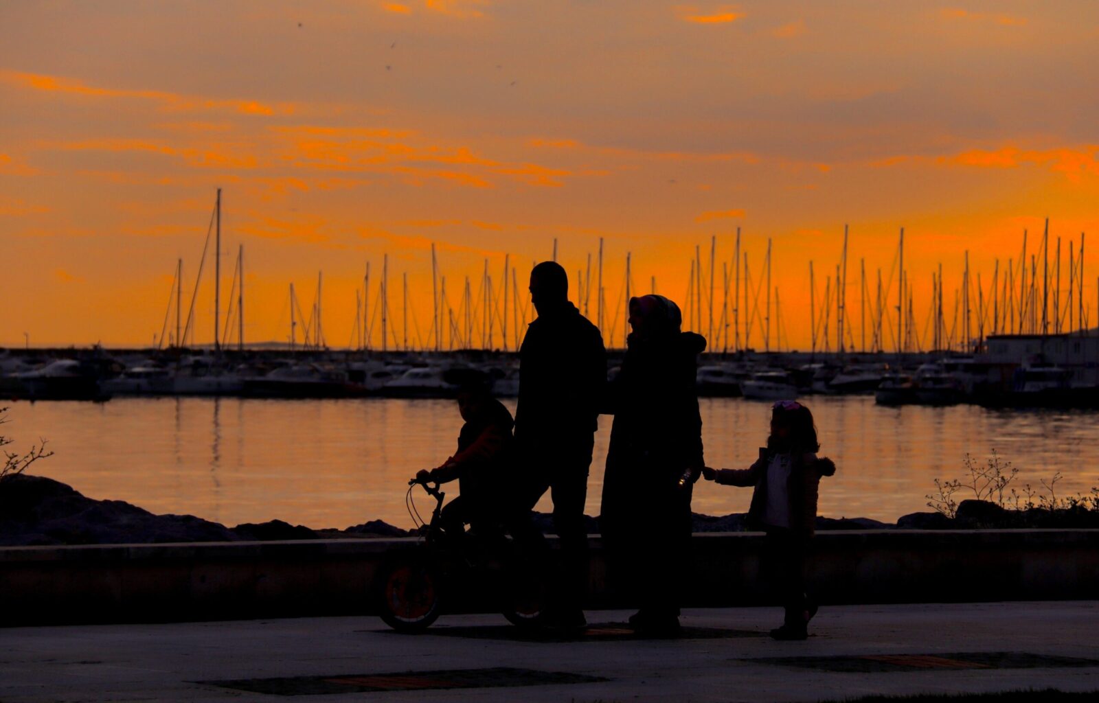 A silhouette of a family walking by the harbor at sunset with boats in the background.