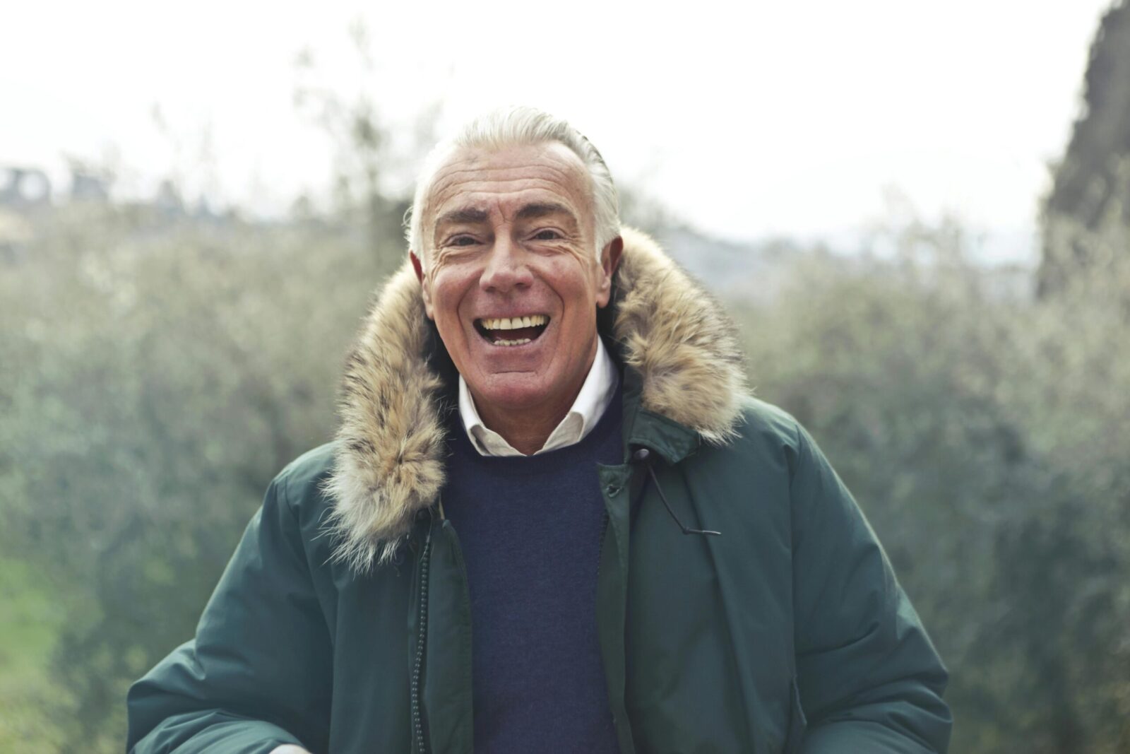 Elderly man with a cheerful smile outdoors wearing a fur-trimmed jacket.