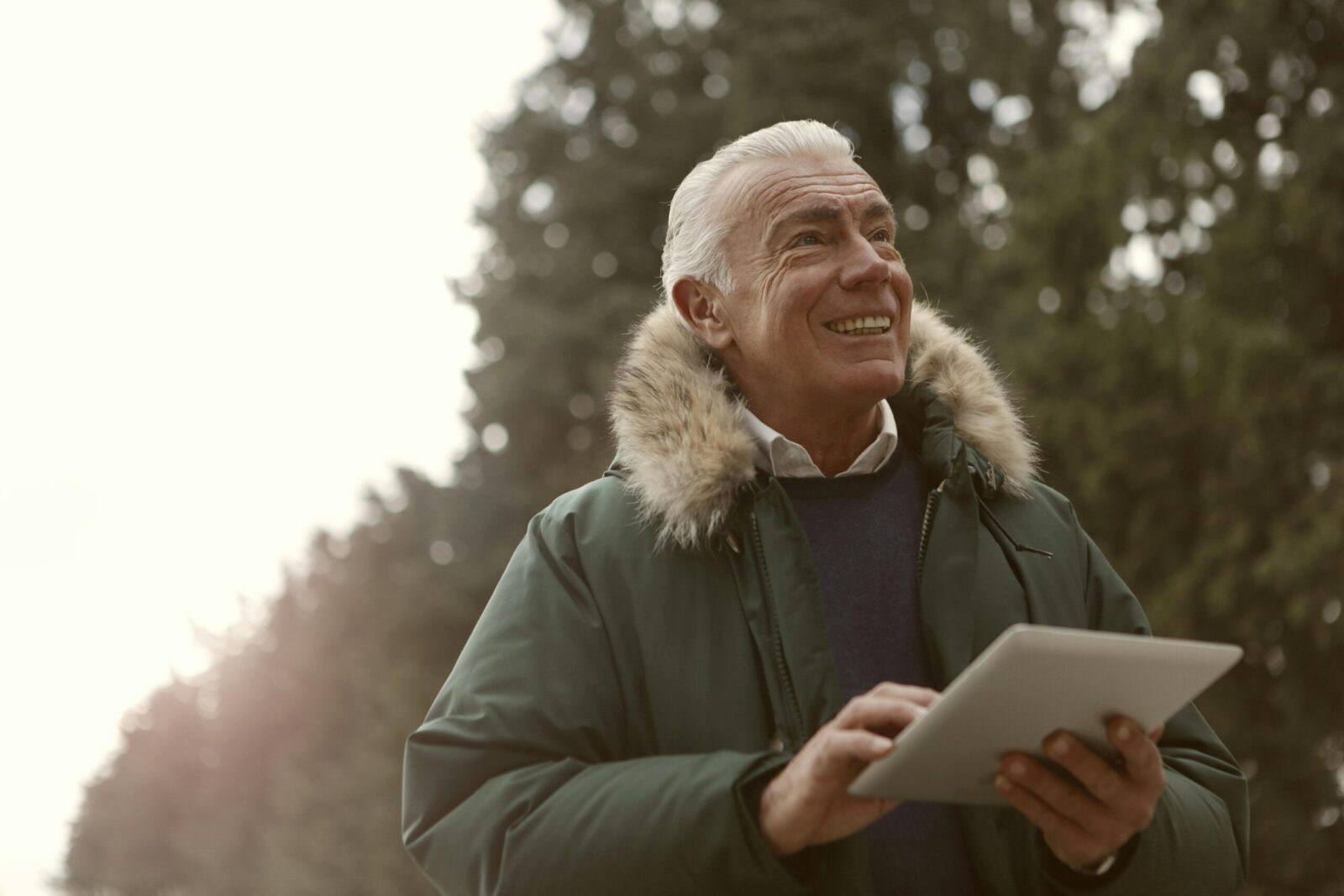 Senior man with tablet smiling outdoors, wearing winter jacket and surrounded by nature.