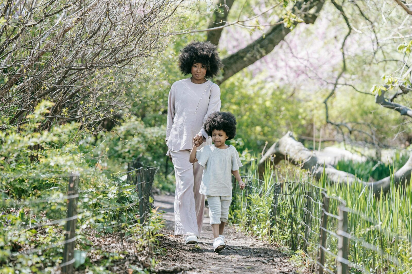 A mother and daughter walk hand in hand along a lush, sunlit path in spring.
