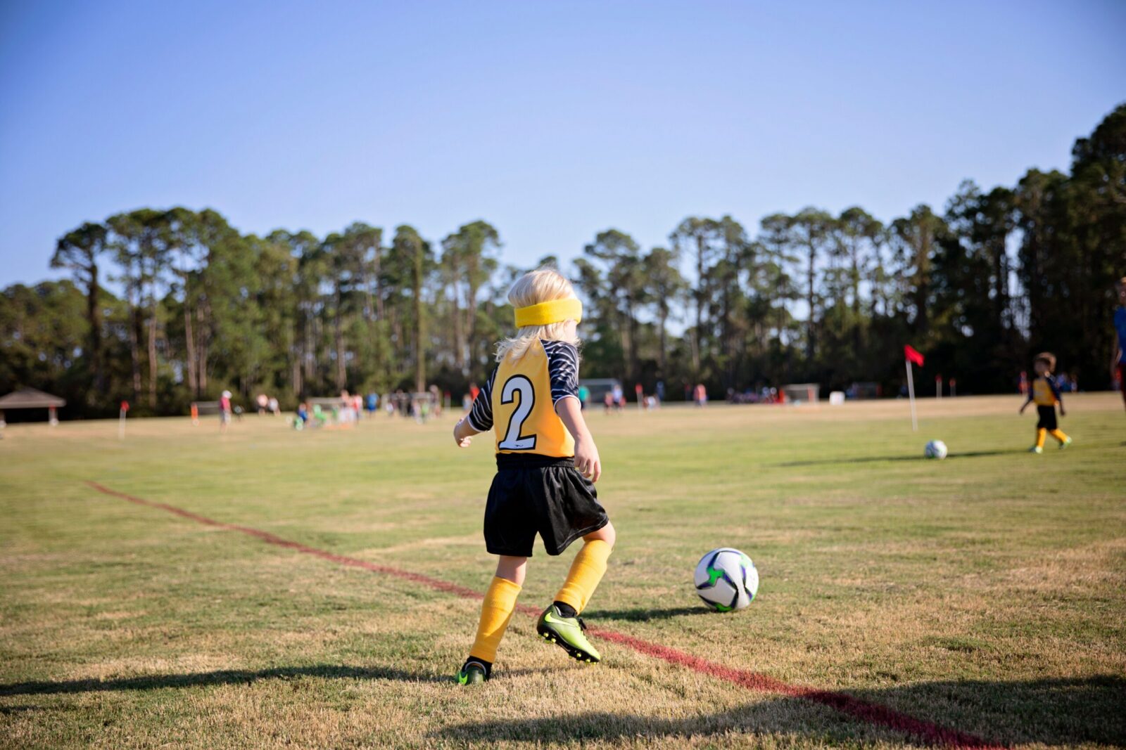 A young soccer player dribbles the ball on a sunny field surrounded by trees.