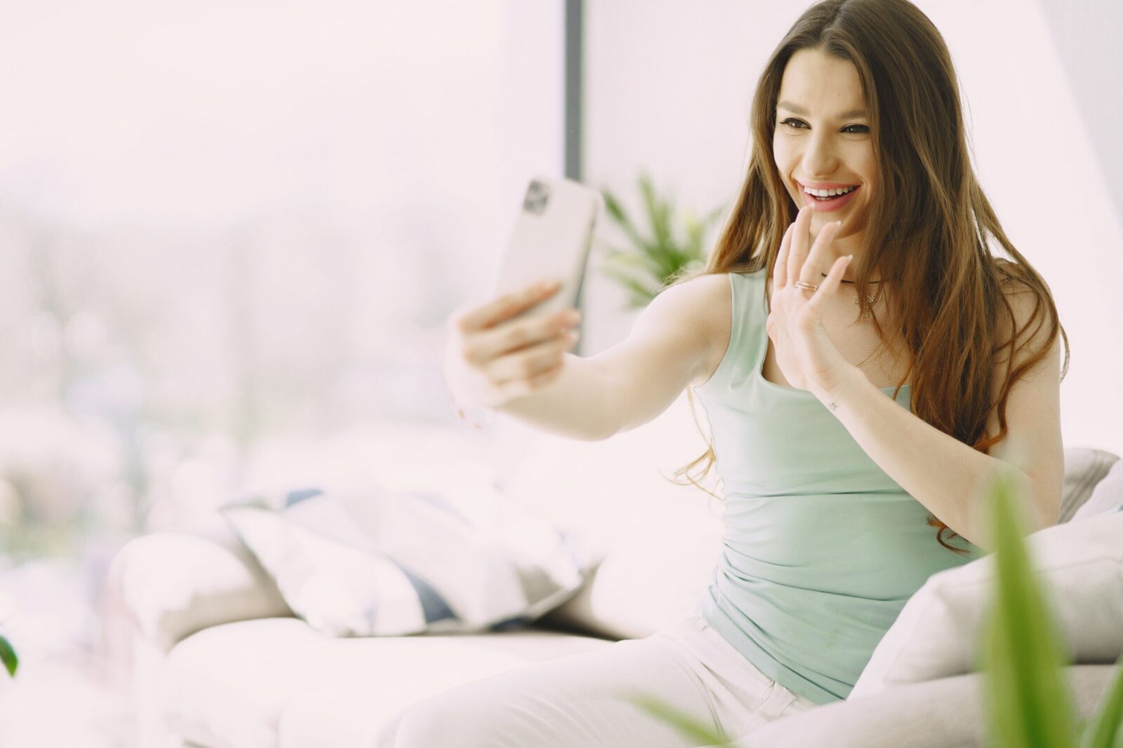 Young happy female in casual clothes greeting with hand and smiling while having video conversation on smartphone in cozy living room