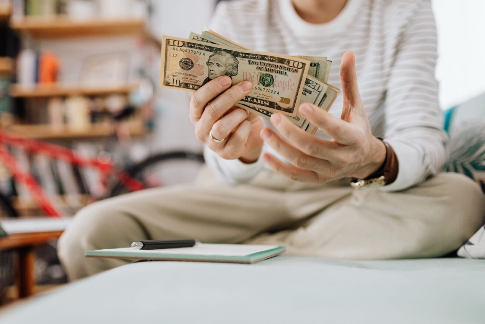 Man wearing casual clothes counts dollar bills while sitting on a sofa indoors.