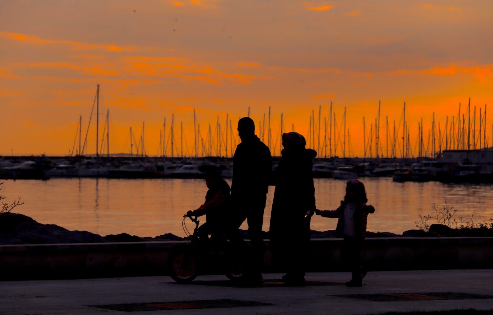 A silhouette of a family walking by the harbor at sunset with boats in the background.